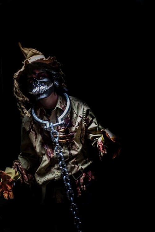A spooky figure in a tattered outfit and skull makeup, wearing a hat and chains, stands against a dark background.