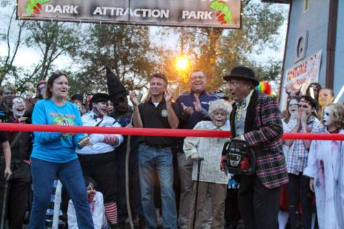 A crowd celebrates at the opening of Dark Attraction Park, with performers in costumes and a man holding a chainsaw.