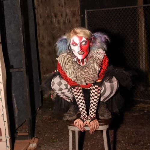 A creepy clown with dramatic makeup and a colorful costume crouches on a stool in a dimly lit outdoor setting.