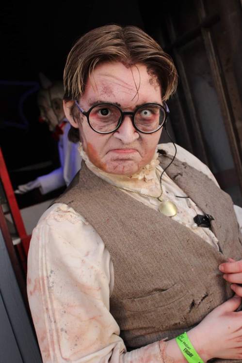 A person in vintage clothing with glasses and a grim expression, featuring makeup for a spooky or horror theme.