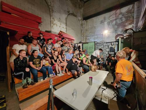 A crowd of people seated in a dimly lit venue, watching an event, with colorful walls and wooden benches.