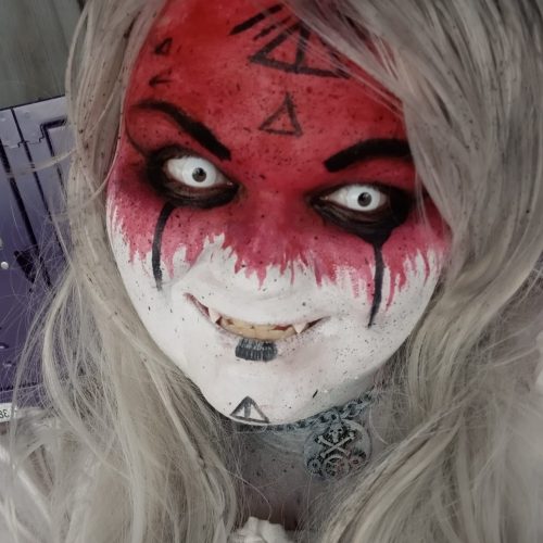 A person with dramatic face paint featuring red and white designs, white eyes, and long white hair, exuding a spooky vibe.
