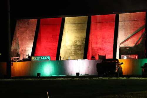 A brightly lit building at night, featuring red, yellow, and green lights, with a white fence and parked vehicles in front.