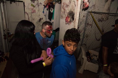 A dimly lit room with a person in a blue hoodie looking back, while another person prepares a balloon. Blood-like effects on the wall.