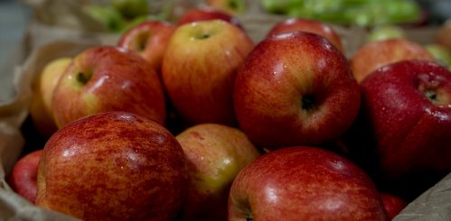 A close-up of a pile of red apples in a brown paper bag, with some green apples blurred in the background.