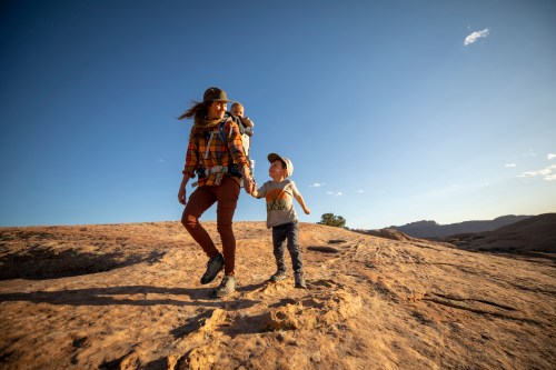 A parent walks on rocky terrain with a child holding their hand and another child in a backpack carrier.