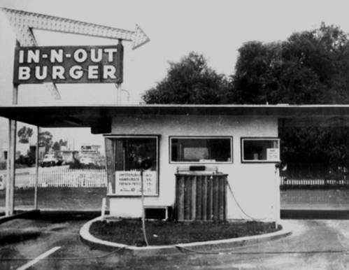 Black and white image of an In-N-Out Burger drive-thru with a sign and a small building.
