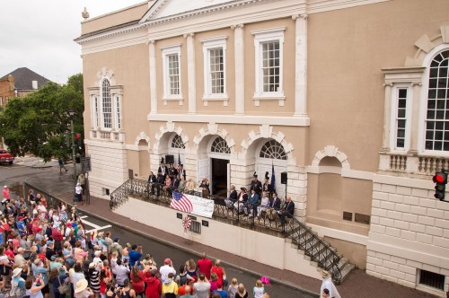A crowd gathers outside a historic building, listening to speakers on a balcony, with an American flag displayed.