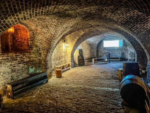 A dimly lit brick cellar with arched ceilings, wooden benches, and barrels, creating a rustic atmosphere.