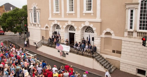 A crowd gathers outside a historic building for a ceremony, with speakers on a balcony and an American flag displayed.
