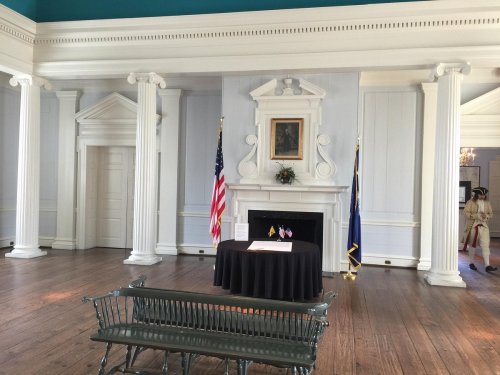 Interior of a historic room with columns, a table displaying documents, and an American flag. A soldier stands in the background.