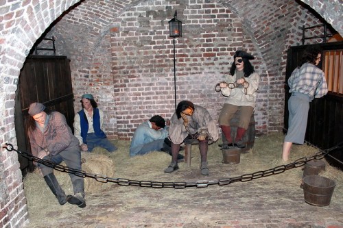 A dimly lit historical dungeon scene with figures in chains, depicting prisoners in a rustic brick setting.