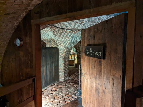 A rustic wooden door labeled "The Provost" opens to a brick archway and a cobblestone floor in a dimly lit space.