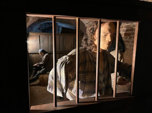A dimly lit scene showing a mannequin behind bars, with a figure sitting in the background, evoking a historical setting.