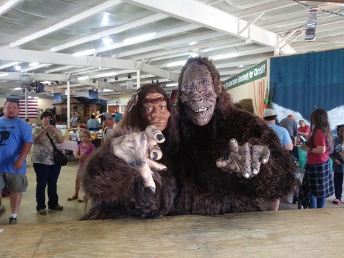 Two people in Sasquatch costumes pose playfully at a fair, surrounded by a crowd of onlookers.