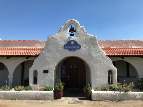 A southwestern-style building with a bell tower and a sign reading "Steamboat," surrounded by potted plants.