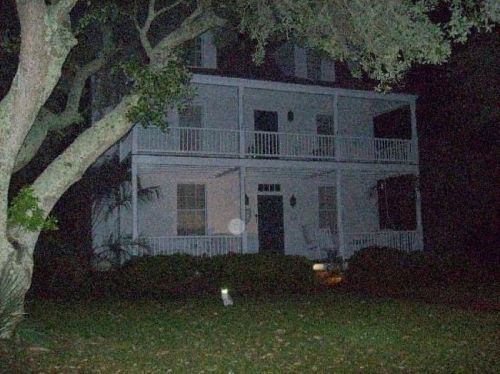 A dark, eerie house with a porch, surrounded by trees, illuminated by a faint light in the foreground.