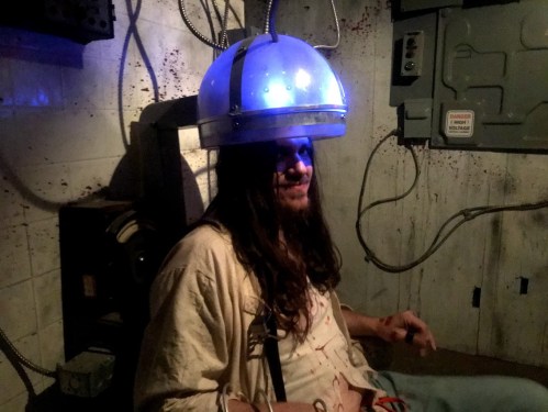A person with long hair sits in a dimly lit room, wearing a futuristic helmet with blue lights, surrounded by eerie decor.