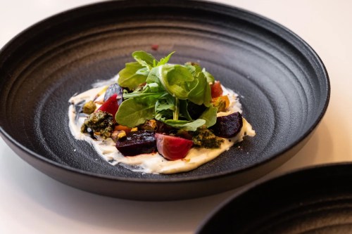 A black plate featuring a colorful salad with beets, cherry tomatoes, greens, and a creamy dressing.