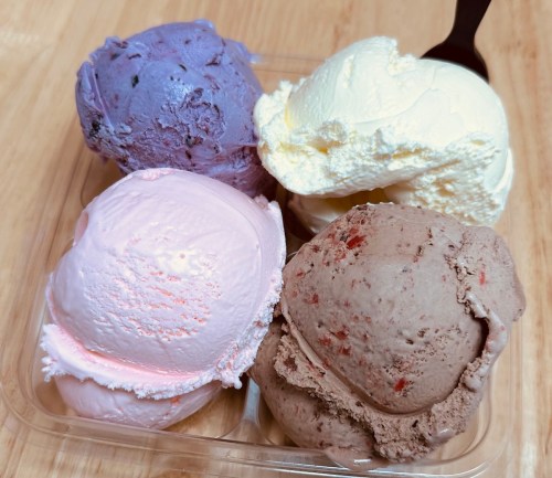 A clear container holds four scoops of colorful ice cream: purple, pink, white, and brown, with a black spoon beside it.