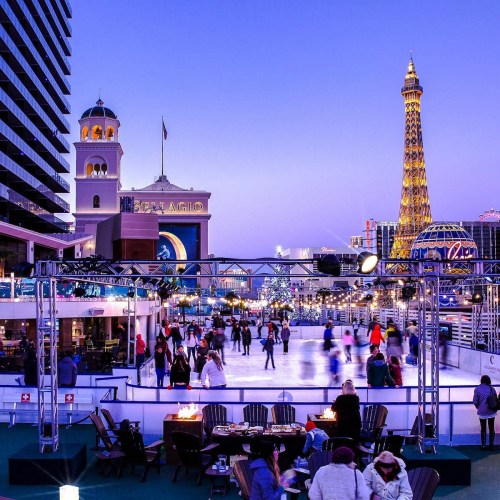 A vibrant outdoor ice skating rink in Las Vegas, with the Eiffel Tower and Bellagio in the background at dusk.