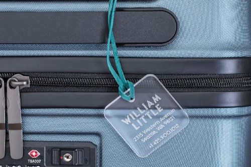 A close-up of a blue suitcase with a clear luggage tag displaying the name "William Little" and contact information.