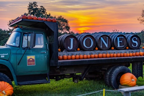 A vintage truck with pumpkins and barrels, set against a colorful sunset. The truck displays the name "JONES."