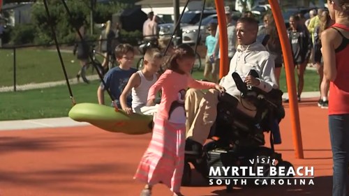 A child plays on a swing while another child approaches a person in a wheelchair at a vibrant park in Myrtle Beach.