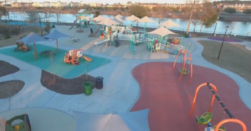 Aerial view of a colorful playground with swings, slides, and shaded areas near a body of water.