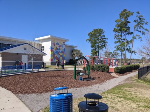 A playground with swings and a colorful building in the background, surrounded by trees and a clear blue sky.
