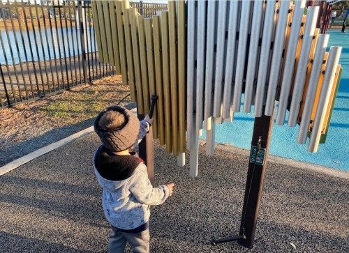 A child in a gray sweater plays with a colorful outdoor musical instrument at a park.