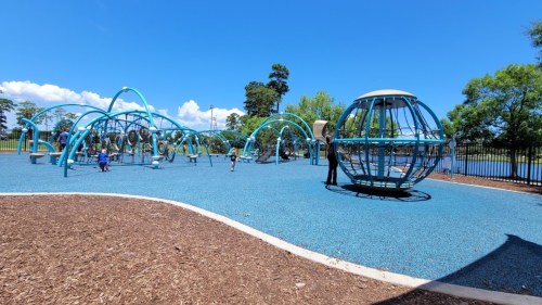 A colorful playground with blue equipment and children playing on a sunny day, surrounded by trees and a clear sky.