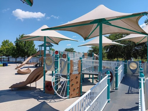 A colorful playground with slides and play structures, shaded by large canopies, set against a clear blue sky.