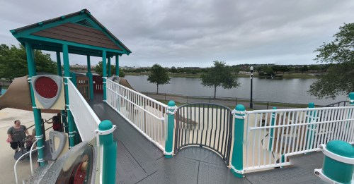 A playground structure with slides and a view of a lake, surrounded by trees under a cloudy sky.