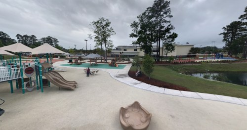A playground with slides, a splash pad, and shaded areas, surrounded by trees and a pond under a cloudy sky.