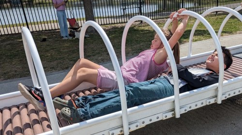 Two children play on a playground structure, one hanging above while the other lies underneath, enjoying a sunny day.