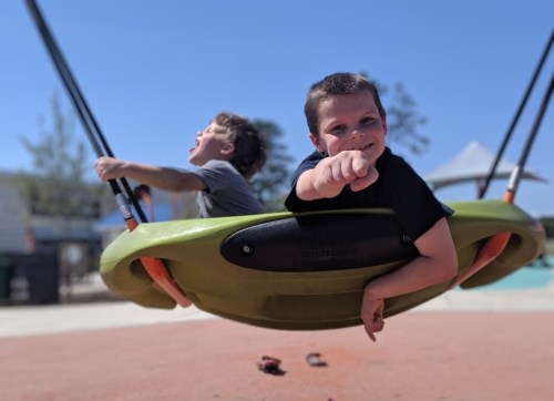 Two boys play on swings at a playground, one smiling and pointing at the camera while the other swings behind him.