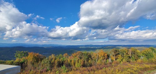 A panoramic view of rolling mountains and colorful autumn trees under a bright blue sky with fluffy clouds.