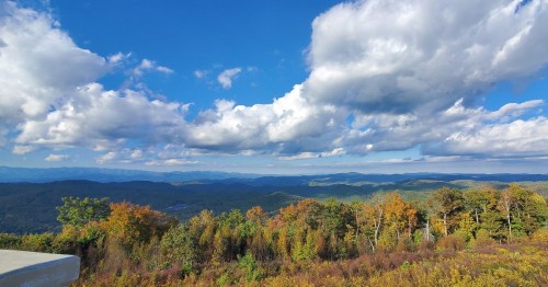 A scenic view of rolling mountains and colorful autumn foliage under a bright blue sky with fluffy clouds.