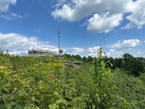 A hillside covered in greenery with a building and communication tower under a blue sky with fluffy clouds.