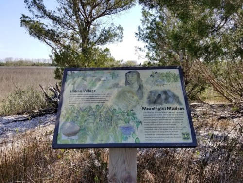 Sign depicting an Indian village and midden, surrounded by tall grass and trees, with a scenic landscape in the background.