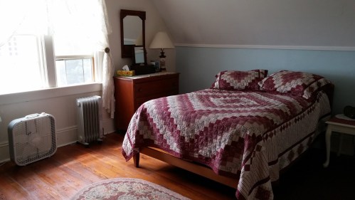 Cozy bedroom with a quilted bed, wooden dresser, fan, and natural light from a window.