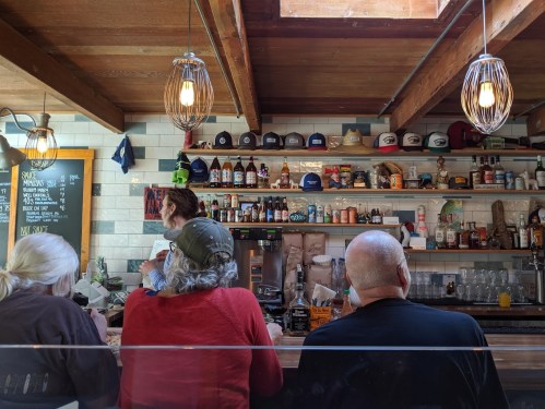 A cozy bar scene with patrons sitting at the counter, a bartender behind, and shelves filled with bottles and hats.