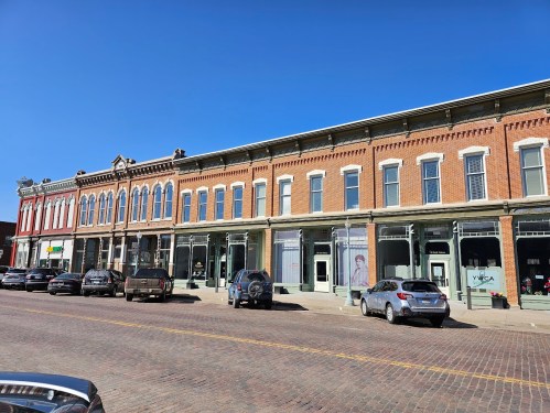 Historic brick buildings line a sunny street, with parked cars and clear blue skies above.