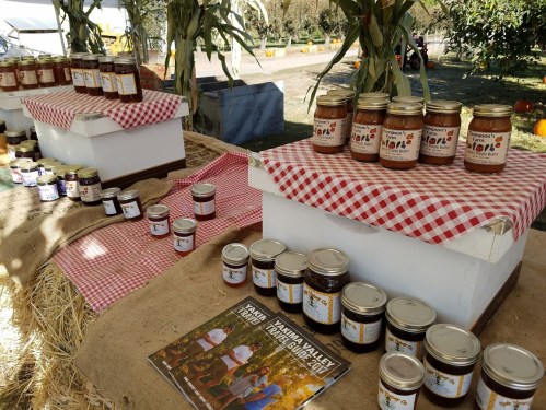 Jars of homemade jams and sauces displayed on tables with checkered cloths at a farm market.