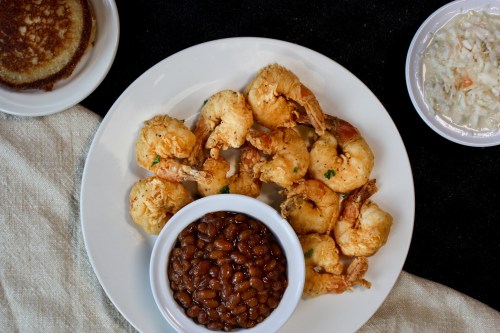 A plate of fried shrimp with a side of baked beans and coleslaw, accompanied by cornbread on a cloth surface.