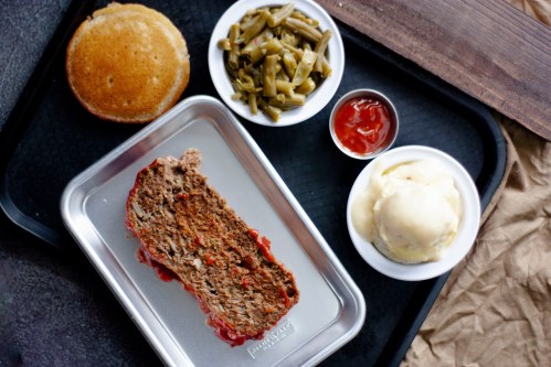 A tray with meatloaf topped with ketchup, green beans, cornbread, and a scoop of mashed potatoes.