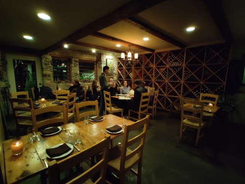 Cozy restaurant interior with wooden tables, chairs, and a wine rack, featuring diners enjoying a meal in warm lighting.