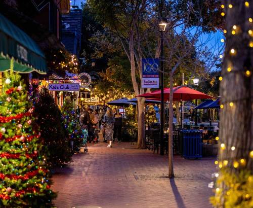 A festive street scene at dusk, adorned with holiday lights, trees, and people enjoying the atmosphere.
