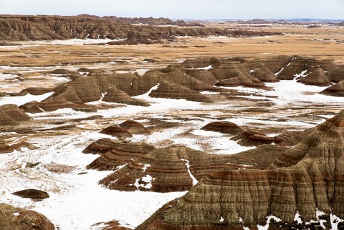A vast landscape of layered hills and valleys, partially covered in snow, under a cloudy sky.